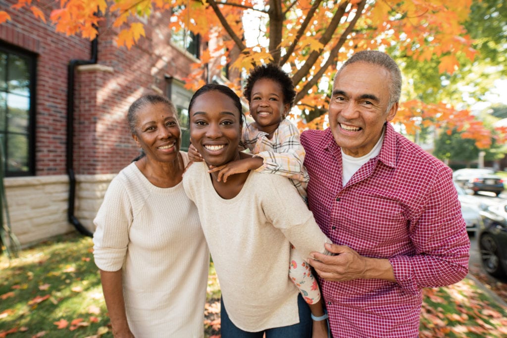 A happy Afro family, it is an image of a mother with her child on her back with the child grandparents beside them. They are outdoors in front of a tree.